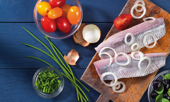 Frische Zutaten für ein Fischgericht auf Holztisch Frischer Fisch mit Zwiebelringen, Tomaten und Schnittlauch auf einem Holzbrett, daneben bunte Tomaten in einer Schüssel und Oliven auf blauem Holztisch