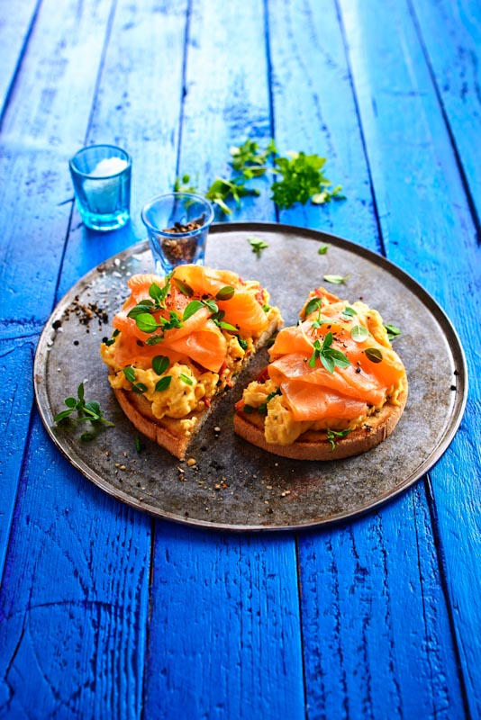 Geröstetes Brot mit Rührei und Lachs, garniert mit Kräutern Zwei Scheiben geröstetes Brot mit cremigem Rührei und zartem Lachs auf einem Teller serviert, dekoriert mit frischen Kräutern.