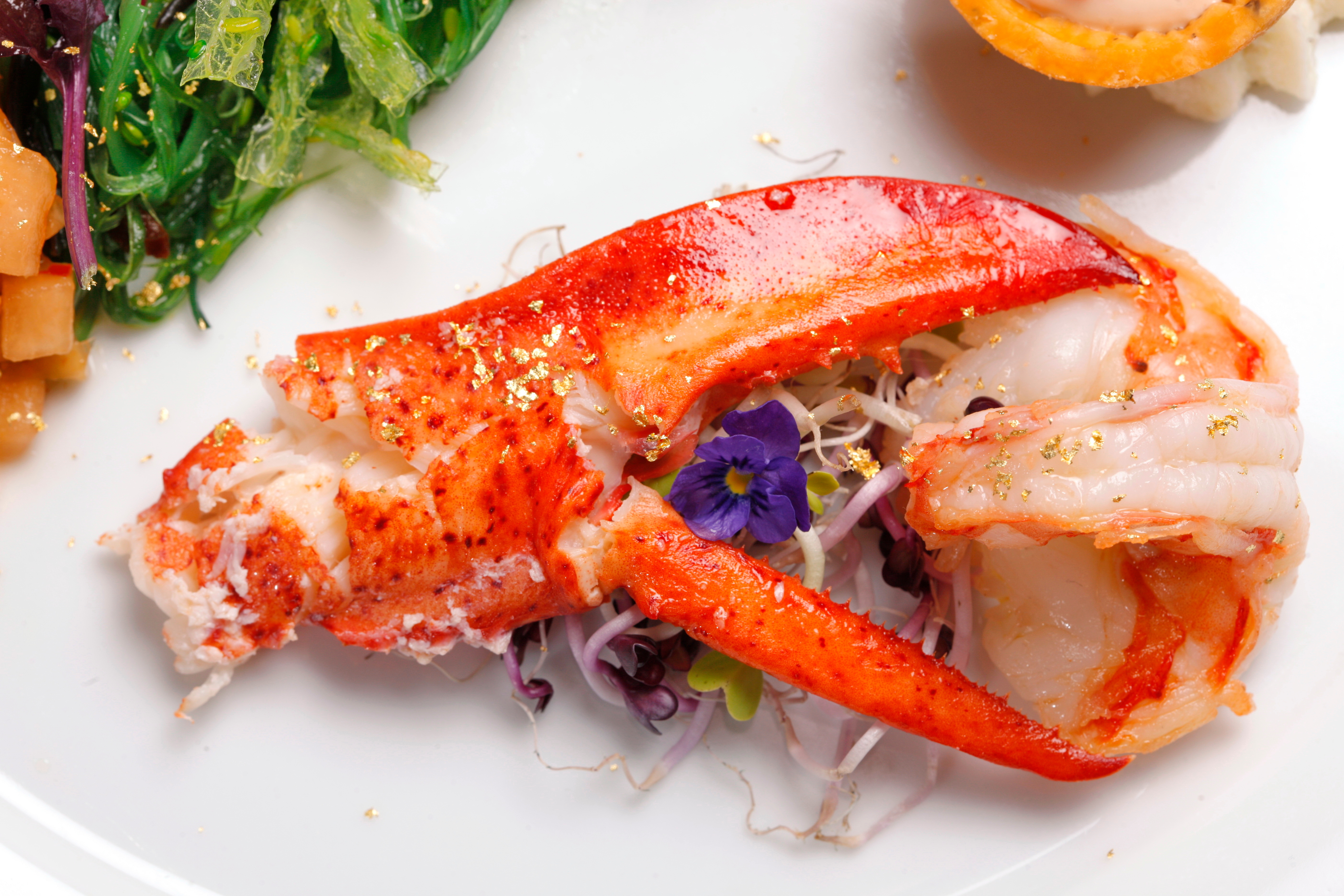 Delikatessen-Teller mit Hummer und essbarem Gold Ein schmackhafter Teller mit einem Hummerstück, garniert mit essbarem Gold, Blumen und Gemüse. Ideal für Gourmets.
