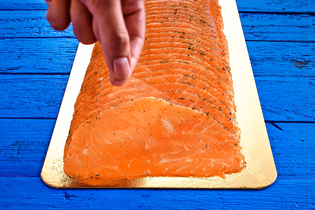 Frische Lachsfilets auf Holzbrett serviert Hand hebt eine Scheibe frischen Lachsfilets von einem Holzbrett auf blauem Hintergrund