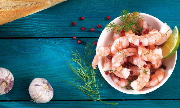 Frischer Garnelensalat mit Dill und Limette Schüssel mit Garnelensalat, garniert mit Dill, roten Pfefferkörnern und Limettenscheibe, auf blauem Holztisch. Knoblauch und Baguette im Hintergrund.
