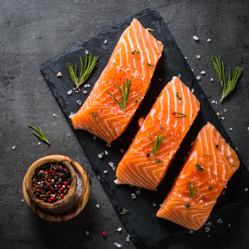 Frische Lachsfilets auf Schieferplatte mit Rosmarin und Pfeffer Drei frische Lachsfilets auf einer Schieferplatte, garniert mit Rosmarin, Salz und Pfefferkörnern, neben einer Holzschale mit buntem Pfeffer.
