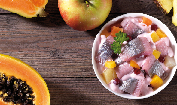 Schüssel mit Heringssalat, Obst und frischem Obst auf Holz Ein Heringssalat in einer weißen Schüssel, umgeben von Apfel, Papaya und anderen Früchten auf einem Holztisch.