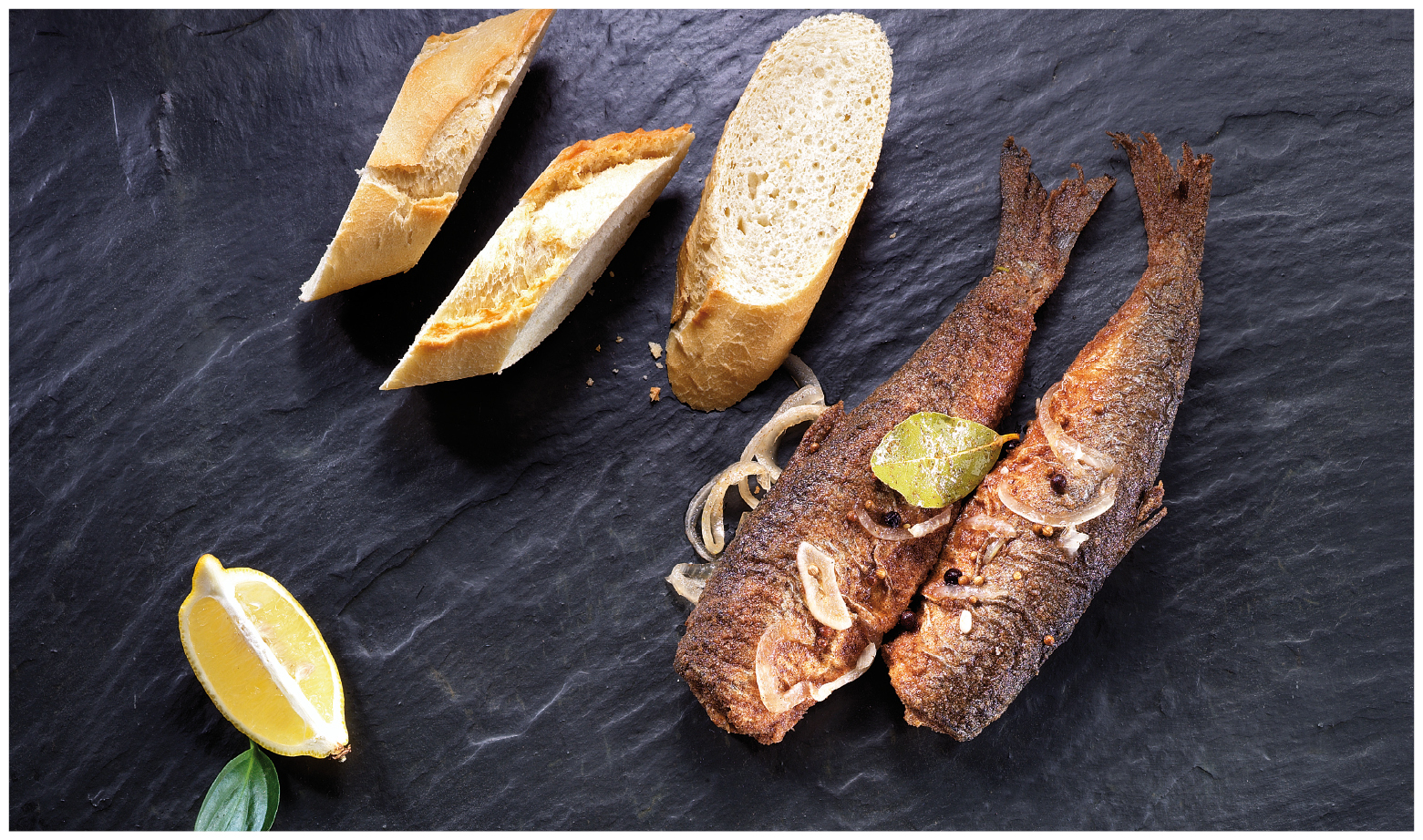 Gebratener Fisch mit Brot und Zitrone auf Schieferplatte Zwei gebratene Fische mit Gewürzen und Limetten auf einer Schieferplatte, serviert mit frischem Baguette und Zitronenscheiben