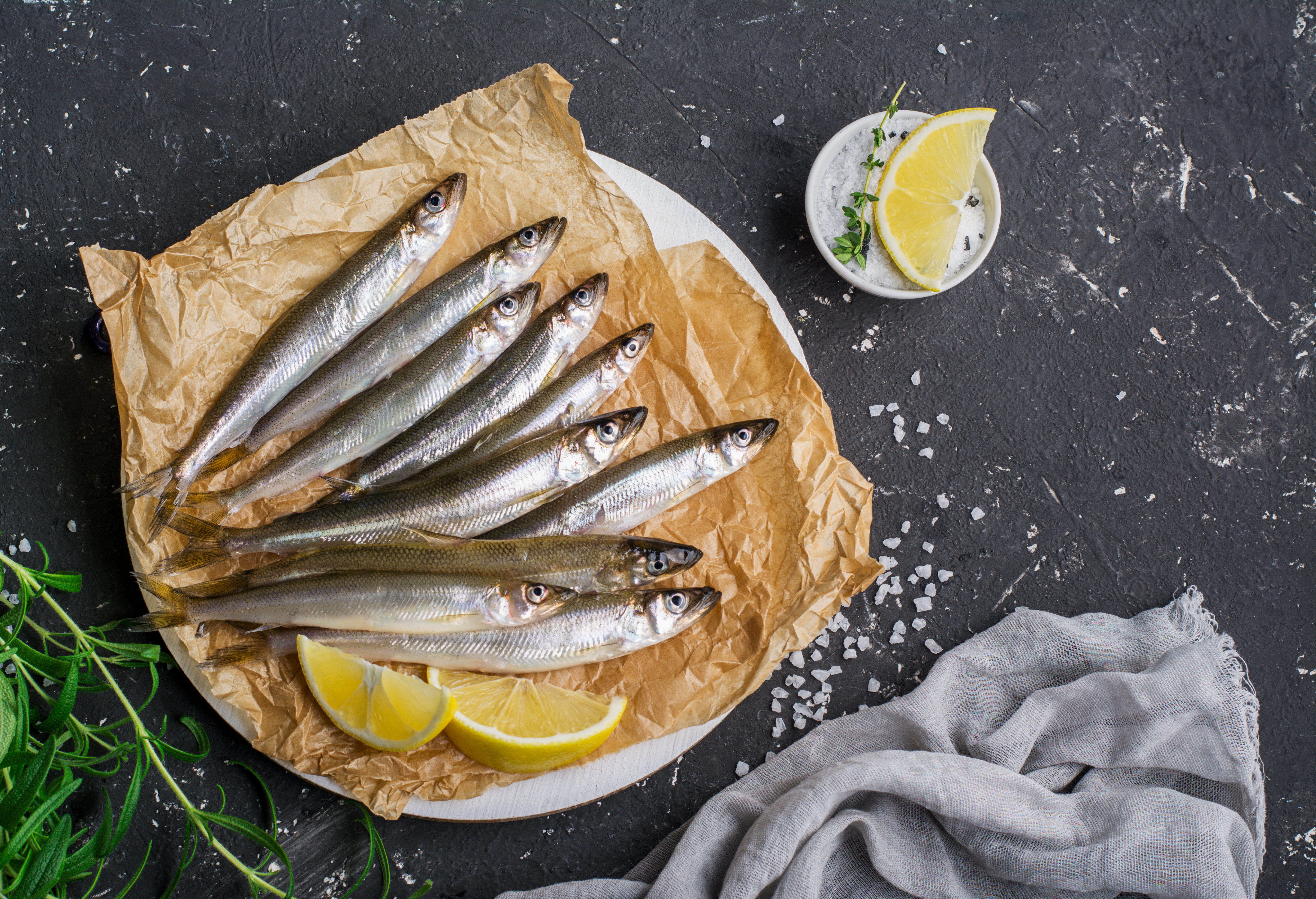 Frische Fische auf Papier mit Zitronen und Salz dekoriert Eine Auswahl frischer Fische auf braunem Papier, mit Zitronenscheiben und Meersalz auf einer dunklen Oberfläche dekoriert. Ein kleiner Becher mit Zitrone und Kräutern ist ebenfalls zu sehen.