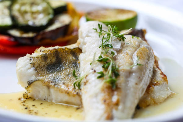 Delikates gebratenes Fischfilet mit Kräutern und Gemüse Nahaufnahme von gebratenem Fischfilet mit frischen Kräutern, serviert mit gegrilltem Gemüse auf einem weißen Teller