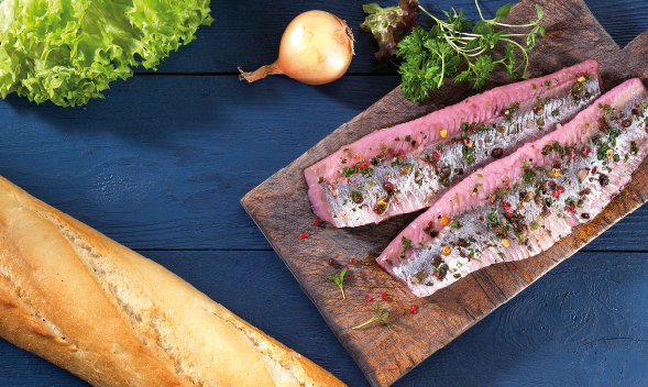 Frische Matjesfilets auf Holzbrett mit Baguette und Gemüsebeilage Zwei Matjesfilets auf einem Holzbrett, garniert mit Kräutern und Pfeffer, daneben ein Baguette, Salat und Zwiebel auf blauem Holzhintergrund