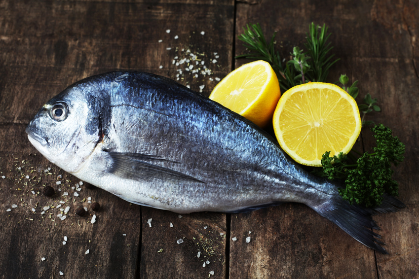 Frischer Fisch mit Zitronen und Kräutern auf Holzbrett Ganze frische Dorade neben geschnittenen Zitronen und Kräutern auf einem rustikalen Holzbrett