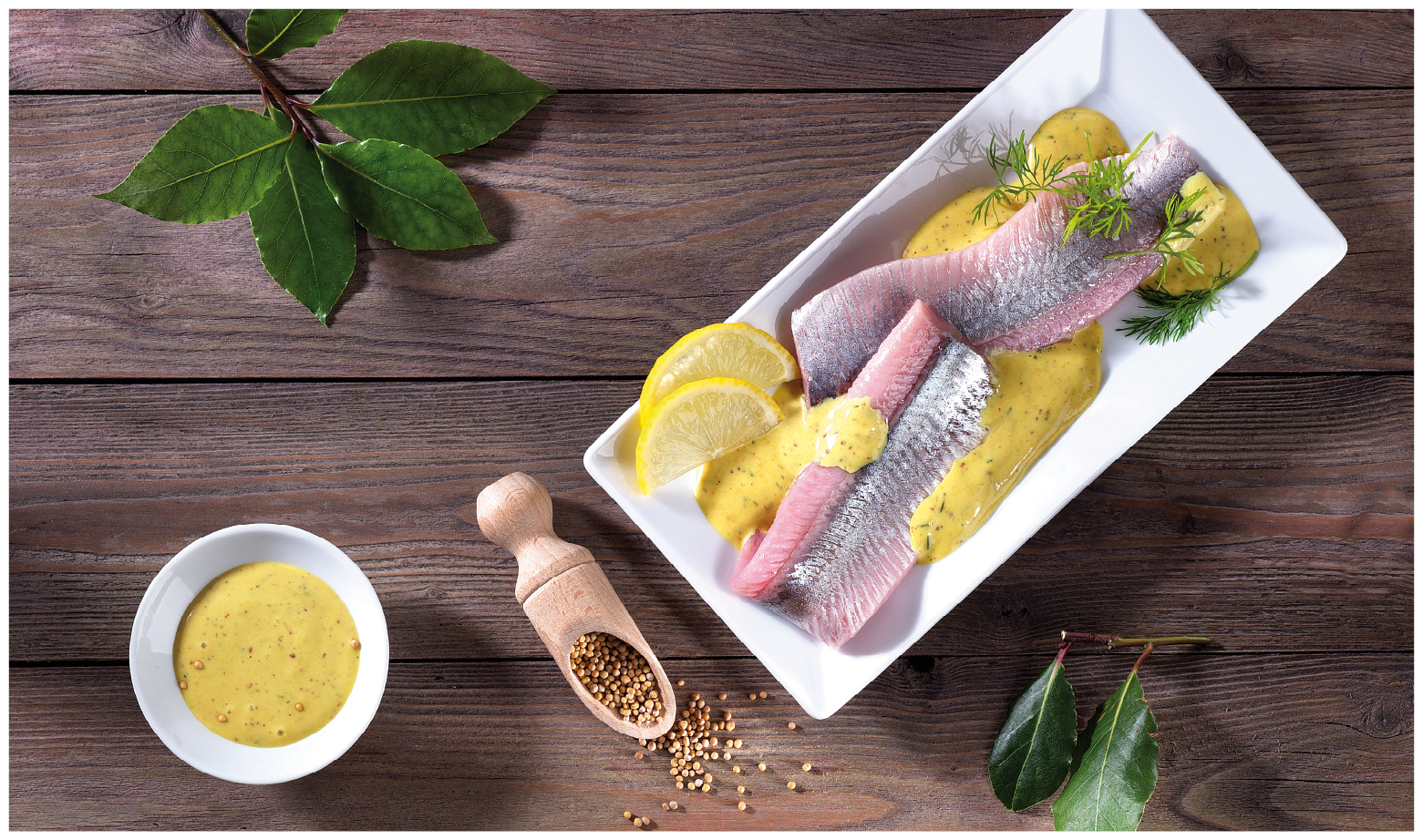 Matjesfilet mit Senfsauce und Zitronenscheiben auf einem weißen Teller Ein Teller mit Matjesfilets, garniert mit Senfsauce, Zitronenscheiben und Dill, auf einem Holztisch. Daneben eine Schale mit Senfsauce und ein Holzlöffel mit Senfkörnern.
