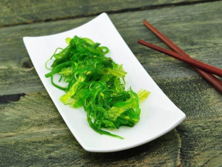 Frischer Wakame-Algensalat auf einem weißen Teller mit Essstäbchen Ein weißer Teller serviert mit frischem grünen Wakame-Algensalat, daneben liegen rote Essstäbchen auf einem Holztisch