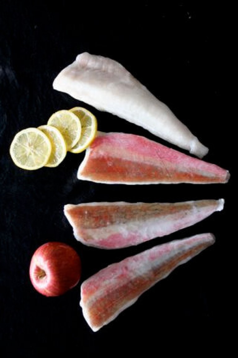Verschiedene Fischfilets mit Zitronenscheiben und Apfel auf schwarzem Hintergrund Drei Arten von Fischfilets, begleitet von Zitronenscheiben und einem roten Apfel, ruhen auf einer dunklen Oberfläche.
