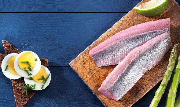 Frische Fischfilets mit Ei, Brot und Spargel serviert Zwei frische Fischfilets auf Holzbrett, daneben gekochtes Ei auf Brot und grüner Spargel