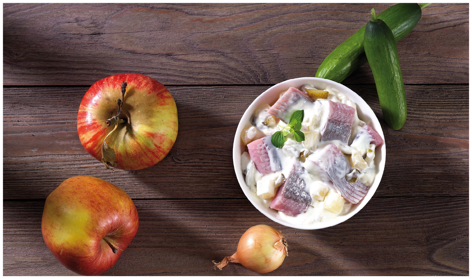 Frischer Heringssalat mit Äpfeln, Gurken und Zwiebeln Schale mit Heringssalat, daneben frische Äpfel, Gurke und Zwiebel auf Holztisch