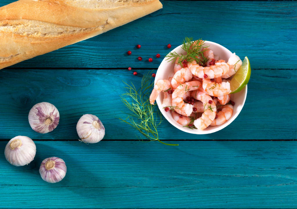 Frische Garnelen mit Kräutern und Limette, Baguette und Knoblauch auf Holztisch Eine Schale mit Garnelen, garniert mit Dill und rosa Pfefferkörnern, Limettenscheibe, daneben Baguette und Knoblauch auf blauem Holztisch